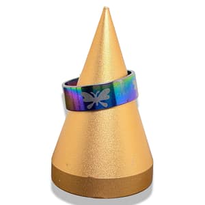 The Anillo Ægir is a metallic, rainbow-coloured ring with butterfly engravings, shown on a gold cone-shaped stand against a white background.